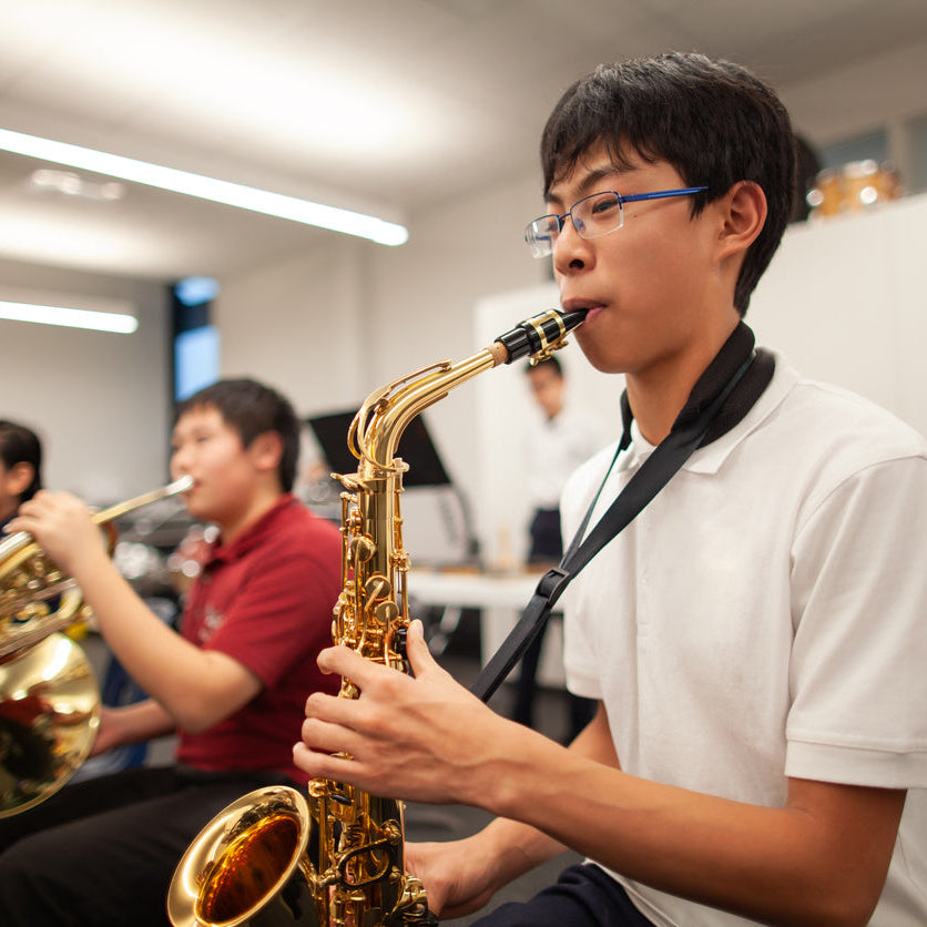 Person playing a saxophone in a music classroom with other musicians in the background.